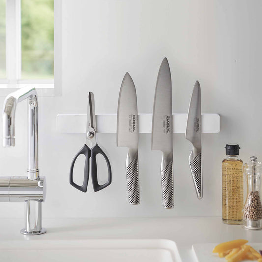 White magnetic knife rack holding kitchen scissors and three knives above a sink.