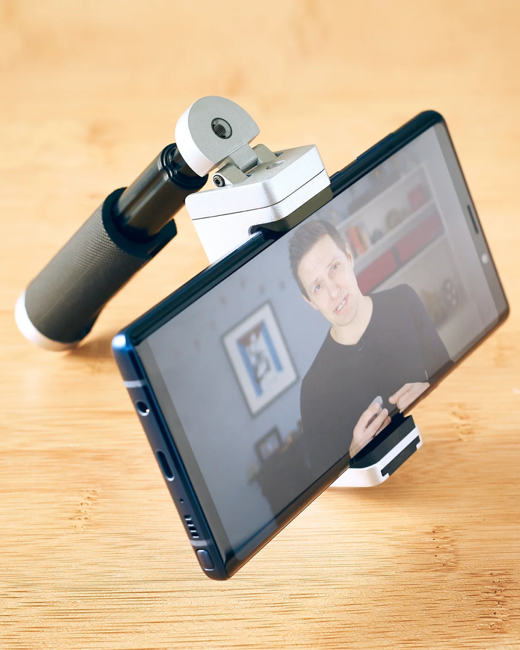 Smartphone mounted on a phone holder with a person on a video call displayed on the screen, on a wooden surface. | Matte Black