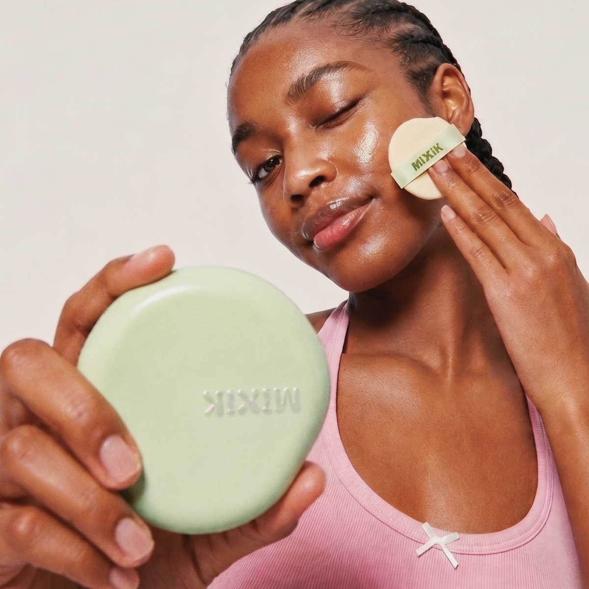 A woman applies a light green MIXIK pressed powder with a puff to her face while holding the round compact in her other hand against a clean off-white background. Cloud Cover Soothing Cushion | Golden Deep