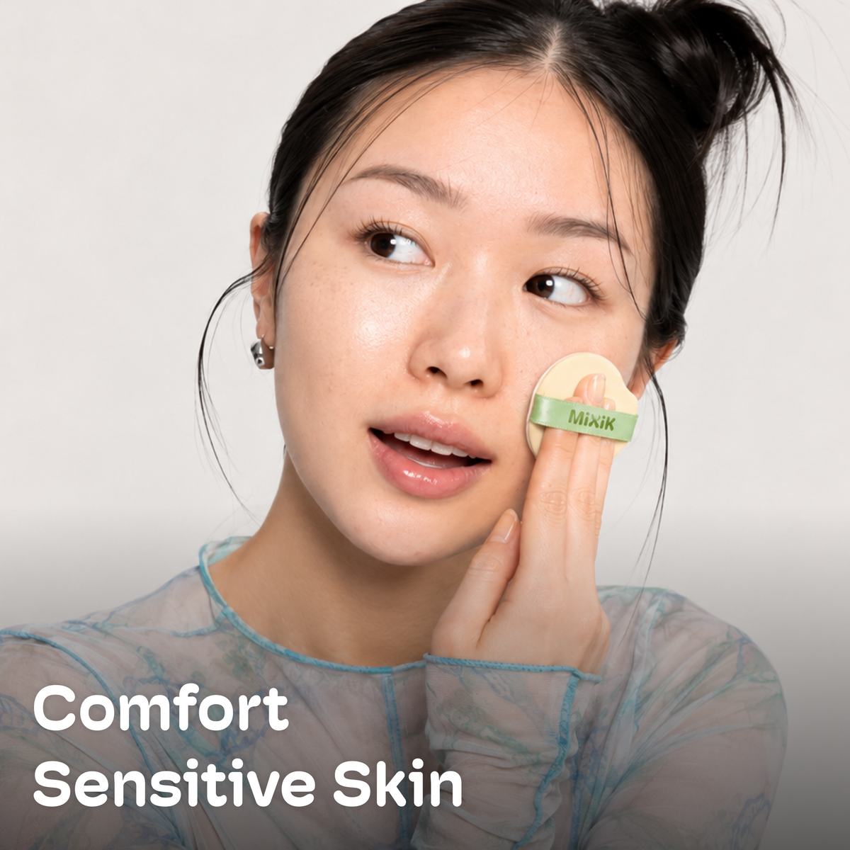 A woman with clear, comfortable skin holds a small beige makeup puff with a green band labeled 'Miwak' against her cheek. White text in the lower left reads 'Comfort Sensitive Skin' on a light gray background. Cloud Cover Soothing Cushion | Golden Medium