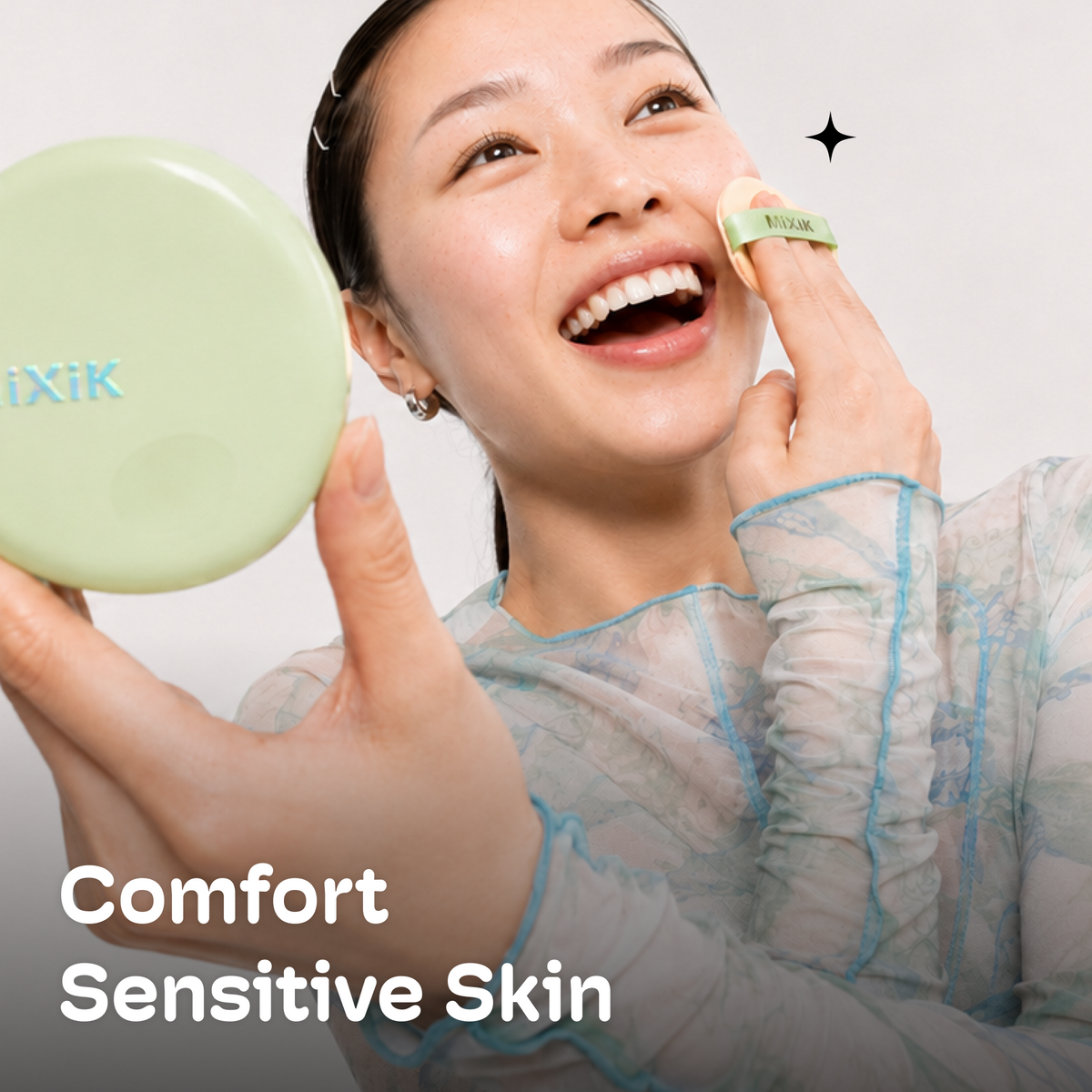 A woman with a radiant smile applies powder from a beige puff to her cheek while holding a light green compact mirror. She is wearing a sheer light blue-gray long-sleeve top. Cloud Cover Soothing Cushion | Medium