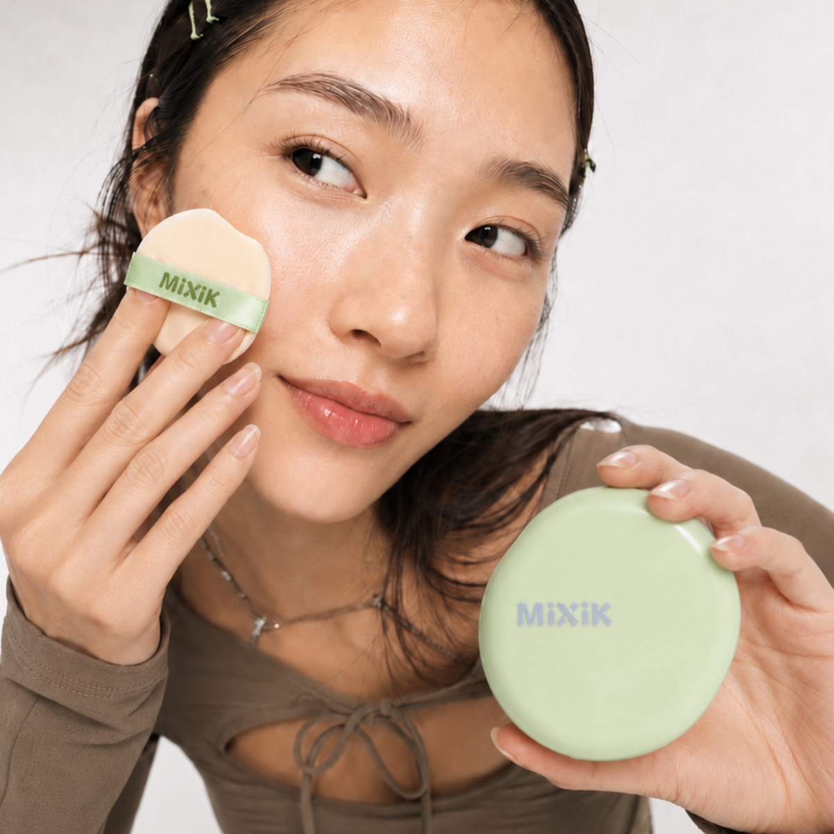 A woman holds a light green round compact and a beige puff with a green band labeled MIXIK, applying makeup against a clean white background. Cloud Cover Soothing Cushion | Natural Medium