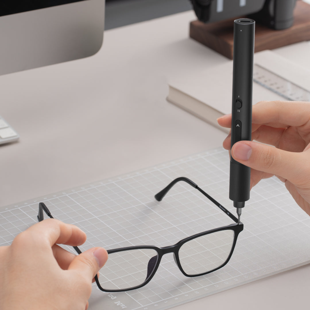 Person using a black tool on a pair of glasses on a desk with a computer and books in the background.