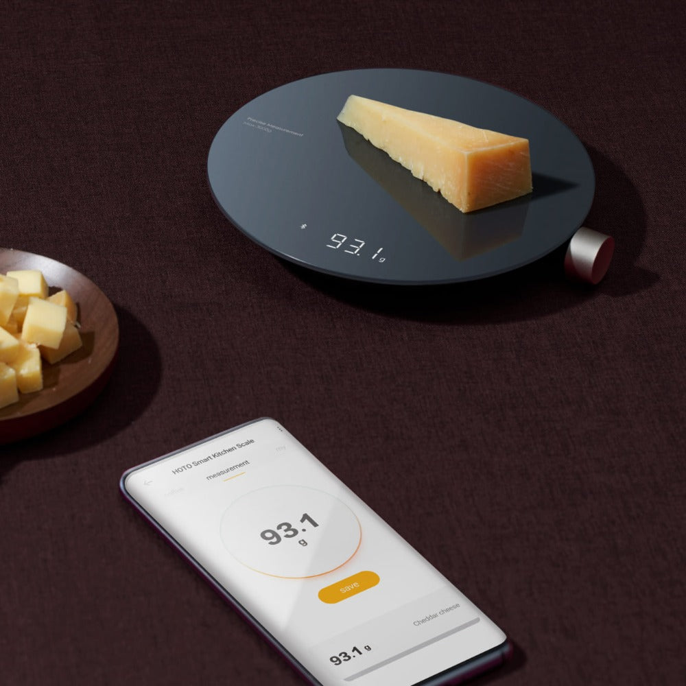 Digital kitchen scale with a piece of cheese on it, next to a smartphone displaying the weight. HOTO Smart Kitchen Scale