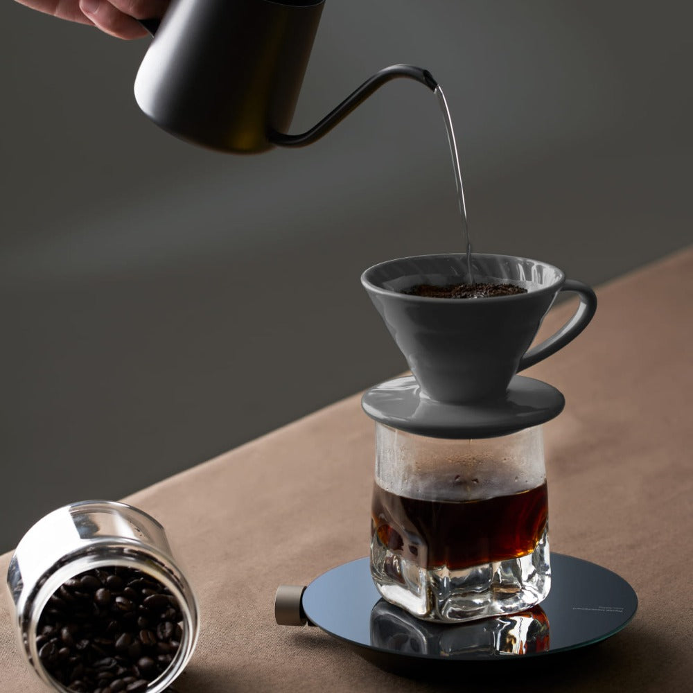 Pouring coffee from a black kettle into a glass dripper on a wooden surface.
