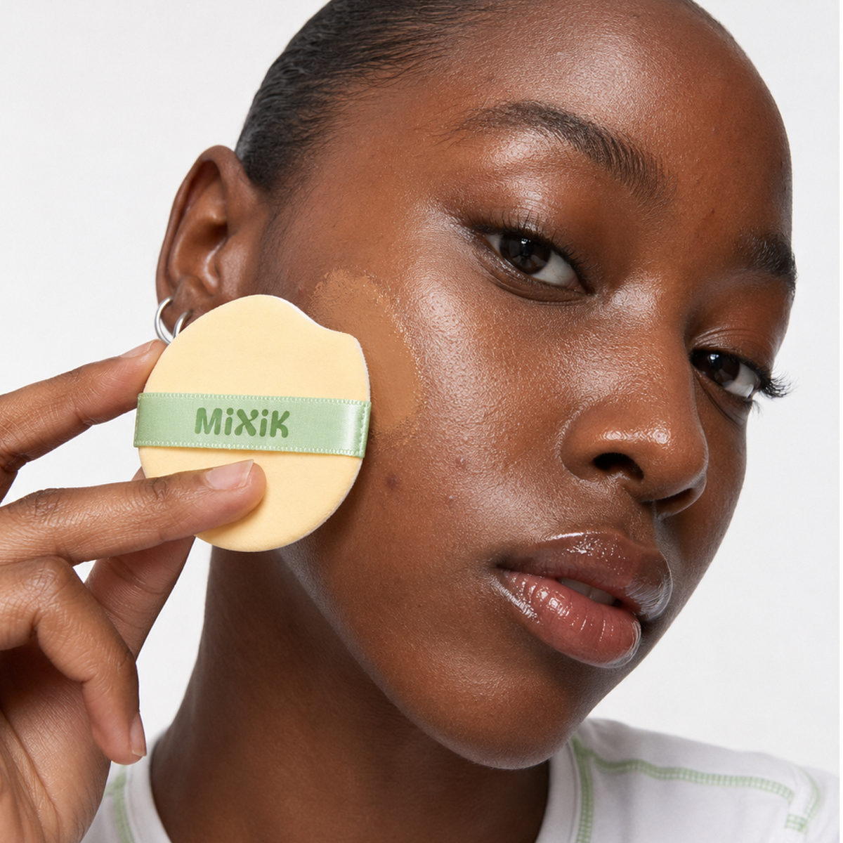 A close-up of a woman applying makeup with a round beige puff sponge featuring a green band with the brand name 'MIXIK' printed on it. MIXIK Cloud Cover Soothing Cushion | Espresso Deep