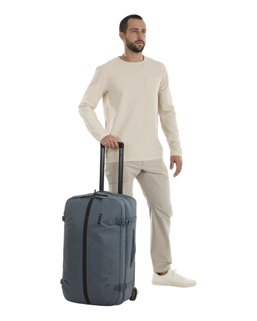 A man wearing a cream long-sleeve shirt, beige pants, and white sneakers stands next to a gray rolling suitcase with black handles and trim on a white background. | Dark Slate