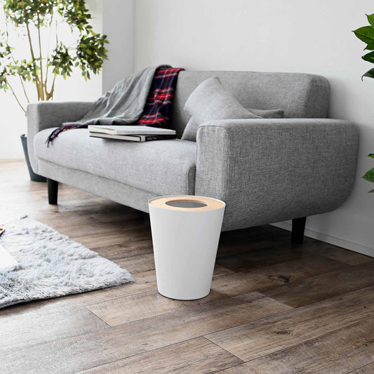 Yamazaki Rin round trash can in the Ash finish, with a white body and wood lid, beside a separate sofa.