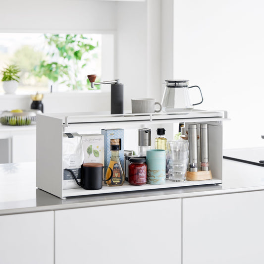 White countertop storage rack with its flap raised, pantry items inside and coffee accessories on top.
