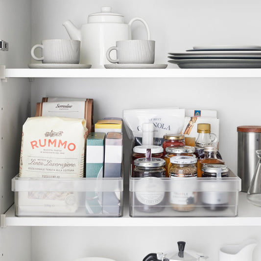 Two translucent organizer bins holding packaged foods and jars on a cupboard shelf; bins sold individually.