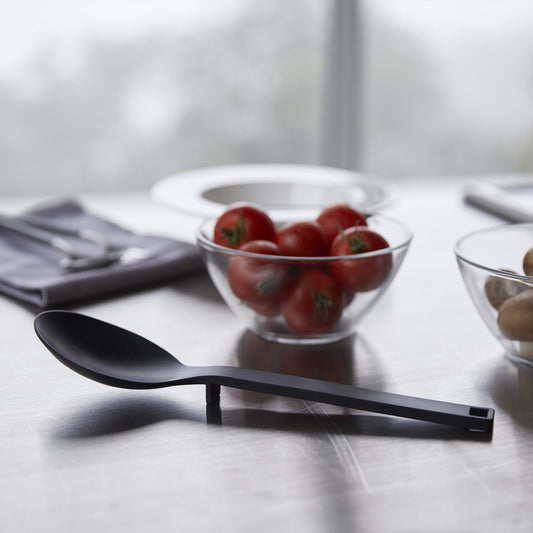 Black Yamazaki Floating Cooking Spoon resting on its built-in support, with the bowl raised above a countertop beside separate ingredients.