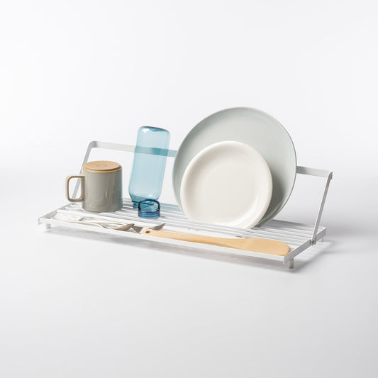 White Yamazaki Slim Over-the-Sink Dish Drying Rack with its support bar raised, shown with separate plates, a bottle, a mug and utensils.