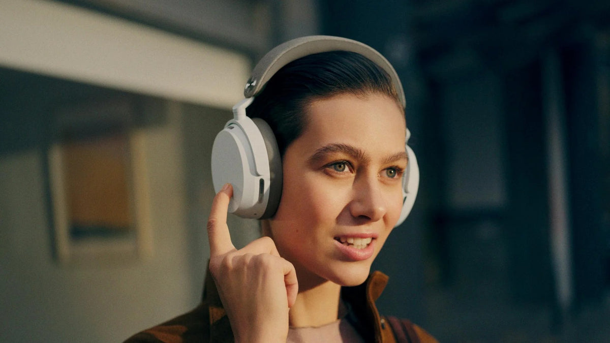 Woman wearing headphones and adjusting them in a room with a blurred background  Sennheiser Momentum 5 Adaptive Noise-Cancelling Around-Ear Bluetooth Wireless Headphone | White
