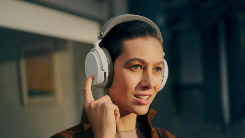 Woman wearing headphones and adjusting them in a room with a blurred background  Sennheiser Momentum 5 Adaptive Noise-Cancelling Around-Ear Bluetooth Wireless Headphone | White
