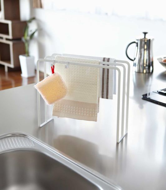 Yamazaki Home Tower Dish Towel Hanger - Steel, white rack drying cloth towels and holding a sponge beside a sink