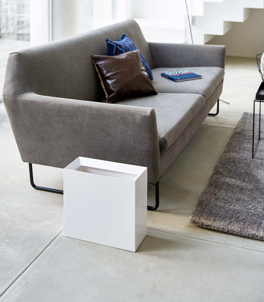 White wide trash can beside a gray sofa in a living room.
